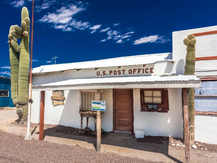 A small white post office that seems to be boarded up; a cactus is in front of it