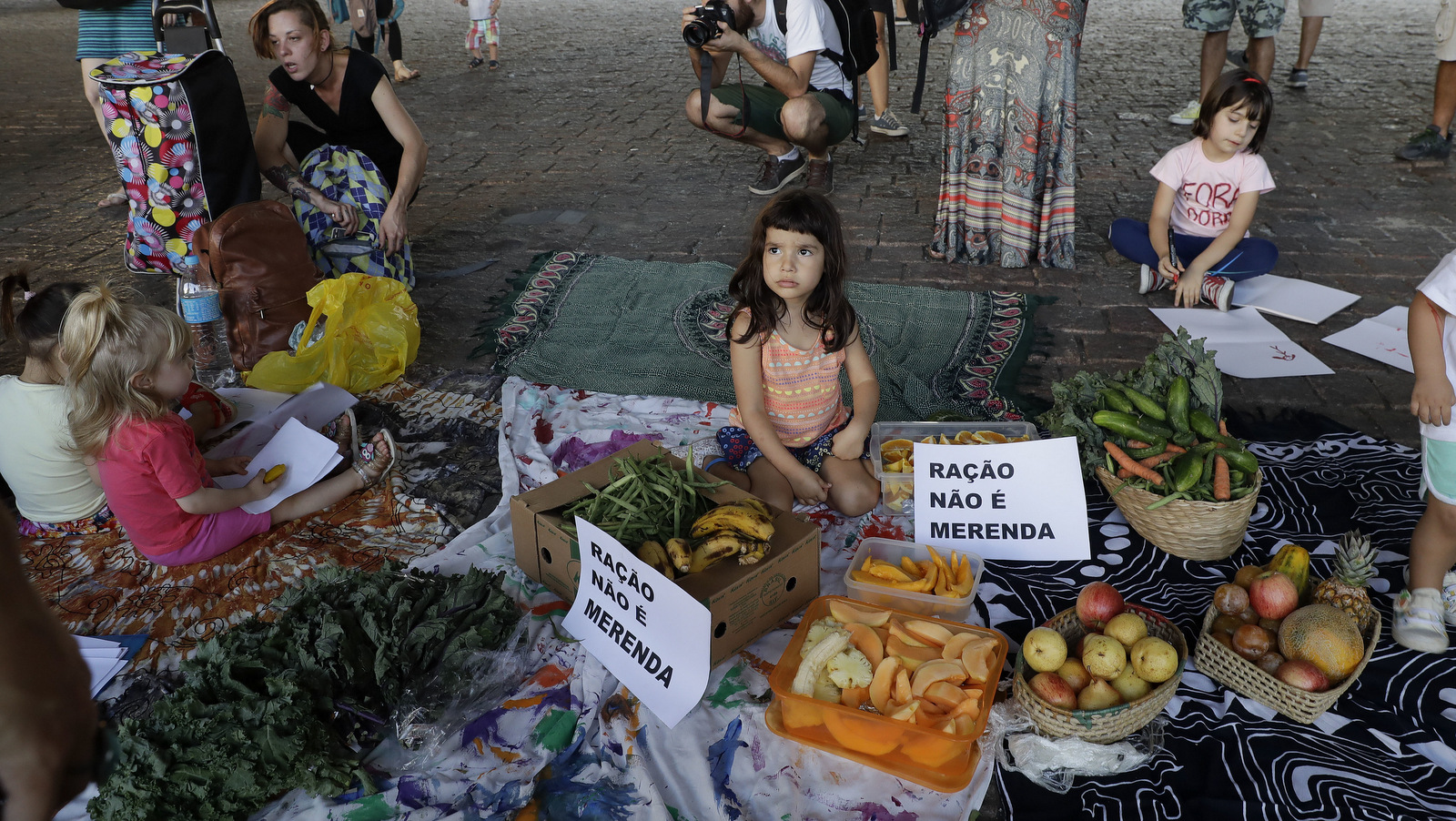 Mothers with their children protest Sao Paulo Mayor Joao Doria's plans to serve school meals made of reprocessed food pellets, next to vegetables and a sign that reads in Portuguese "Pellets are not meals" in Sao Paulo, Brazil, Thursday, Oct. 19, 2017. The pellets are made of dehydrated leftovers and resemble popcorn, and some are mixed into other foods, like cakes, while others can be eaten directly. (AP Photo/Andre Penner)