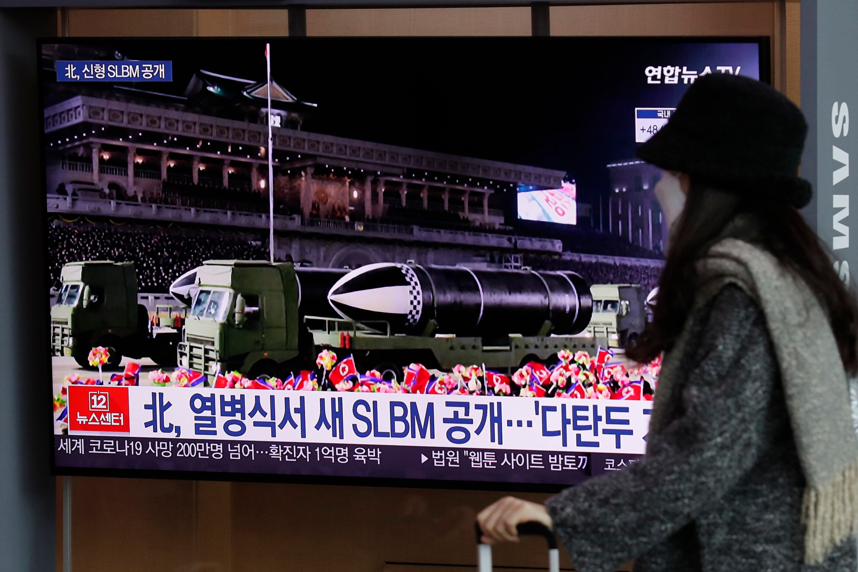 A woman wearing a face mask walks past in front of a TV screen showing a news program reporting about North Korea's military 