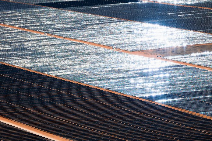 Aerial view of endless rows of solar panels, some gleaming in the sunlight, in a red dirt desert.