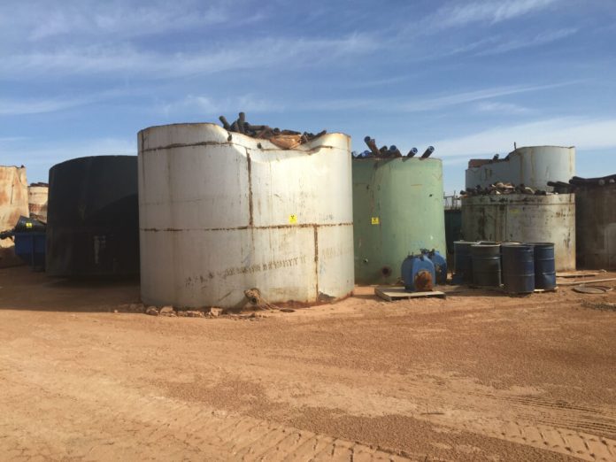 Large, rusty, and damaged white, gray, and green tanks hold oilfield pipe waste, with blue steel drums and dumpster nearby.