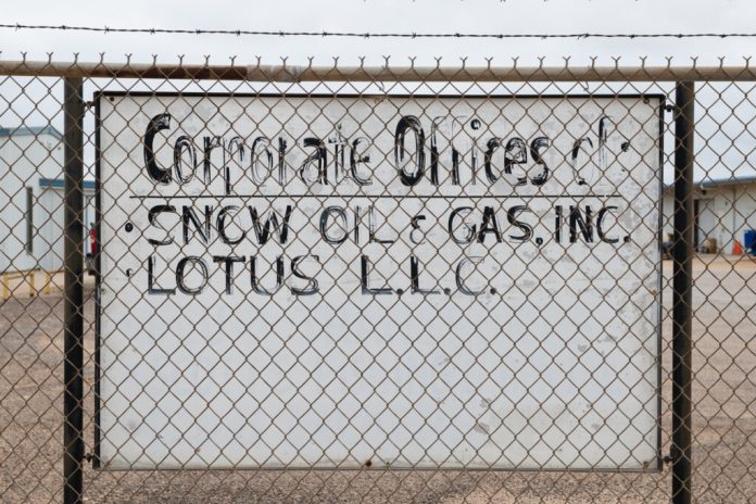 Large white sign with faded black paint with text 'Corporate offices of Snow Oil and Gas Inc, Lotus LLC' behind a chainlink fence