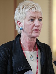Middle-aged woman with short, very-light hair speaking into a microphone.