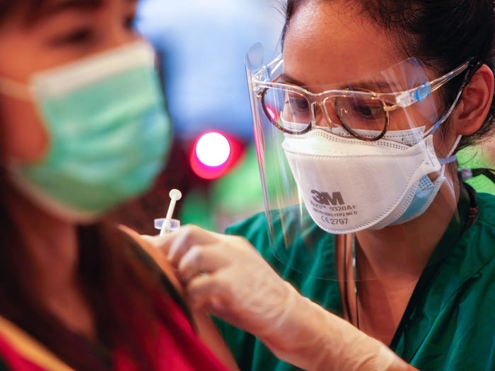 A woman wearing glasses, a mask and a face shield administers a doses of Sinovac Coronavac vaccine to another woman, blurred, in the foreground, who is also wearing a mask, in Thailand in May, 2021.