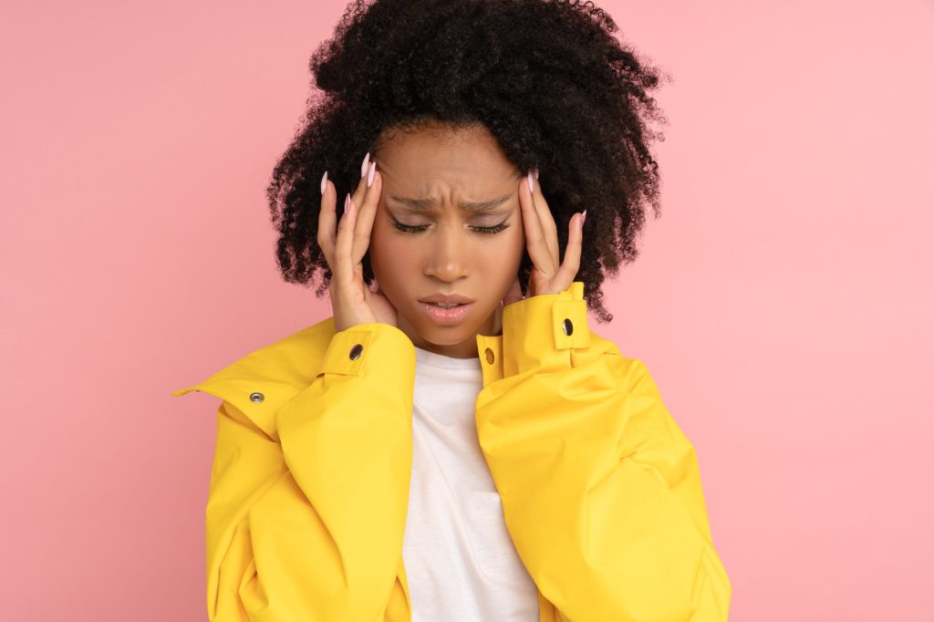 Unhappy Afro woman wear yellow raincoat suffers from severe throbbing headache touching her temples