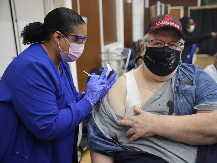 man in a mask getting his third booster shot of Pfizer vaccine injected into his right arm by a healthcare worker (also masked).