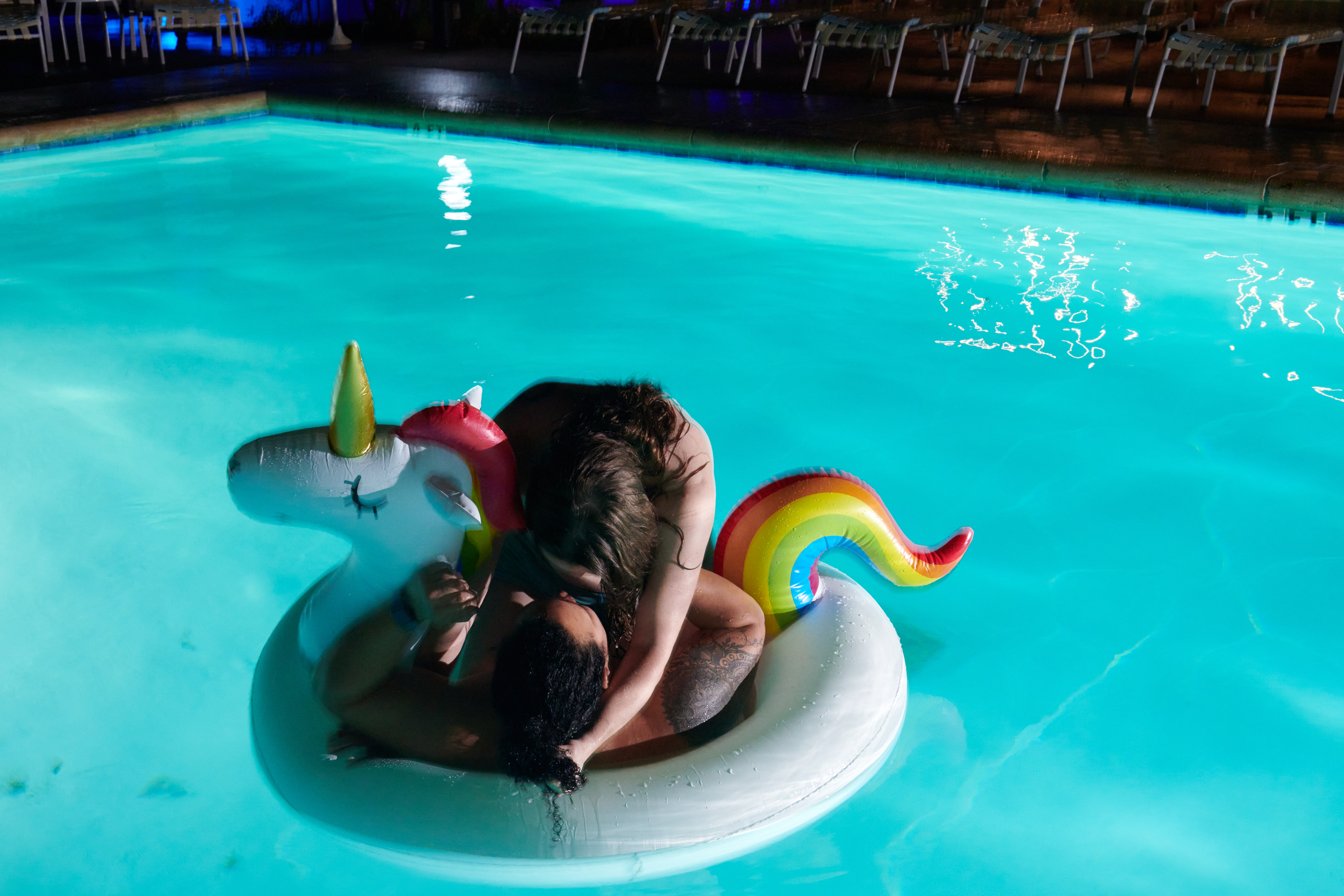 Two nude people kiss on a unicorn floaty in a pool, their faces are obscured.