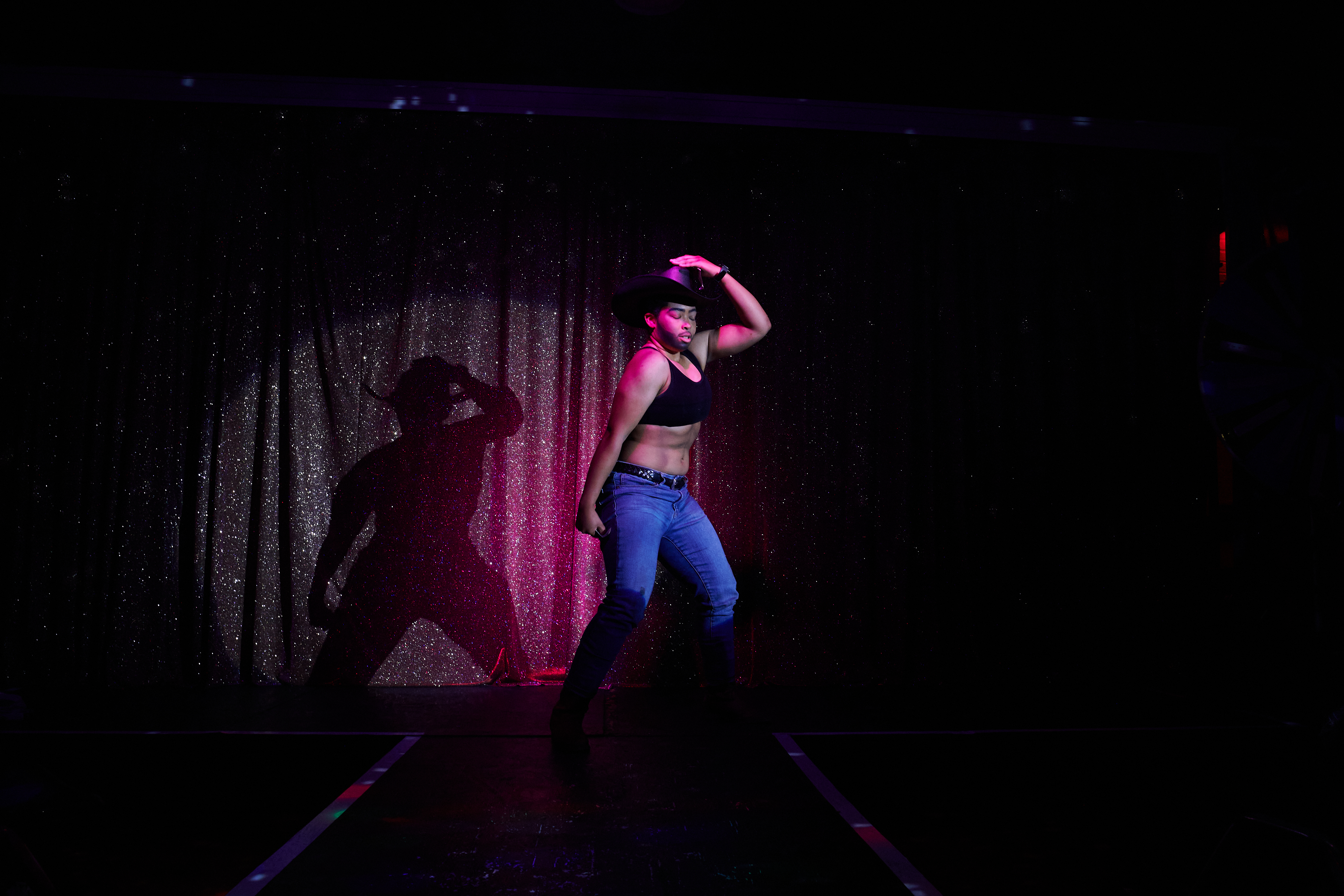 A performer is onstage wearing a sports bra, denim, and a black cowboy hat, striking a pose in front of a spotlight.