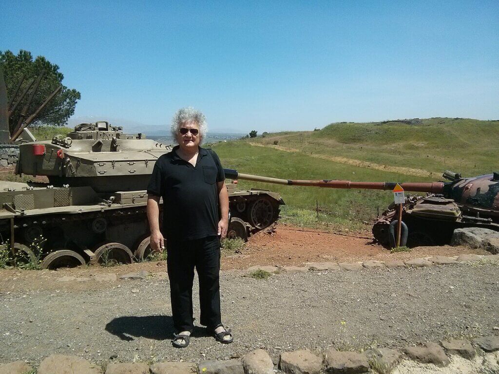 Ephraim Laor standing in the occupied Syrian Golan Heights in April 2014, with two tanks behind him and rolling green hills in the background.